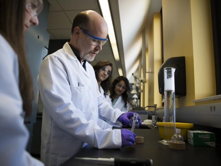 Students in lab coats and safety glasses observe professor measuring chemical reagents in graduated cylinder